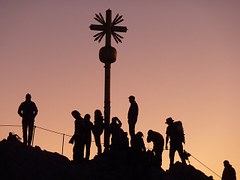 Das Bild zeigt ein Gipfelkreuz im Gegenlicht, mit Menschen drumrum